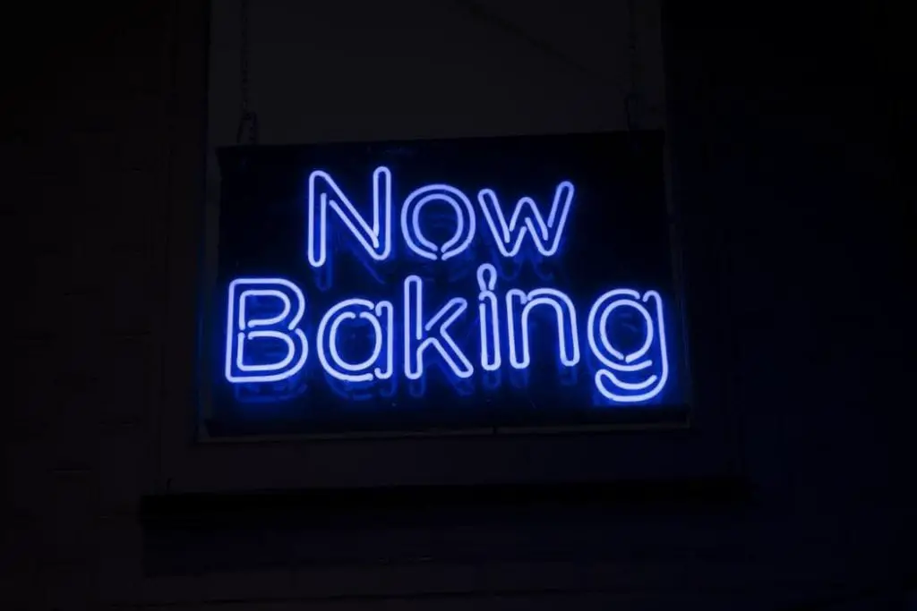 A blue neon sign hanging on a wall that reads ‘Now Baking’.